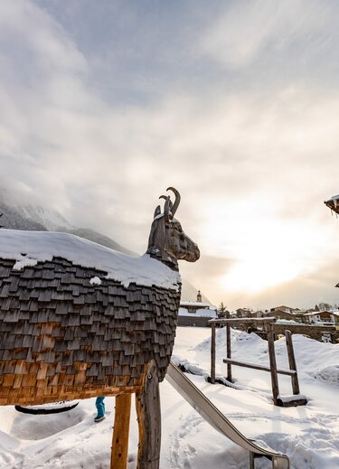 Holzstatue eines Steinbocks mit Schnee auf dem Dach vor verschneiter Berglandschaft