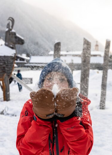 Kind mit Fäustlingen und roter Jacke pustet Schnee in die Luft