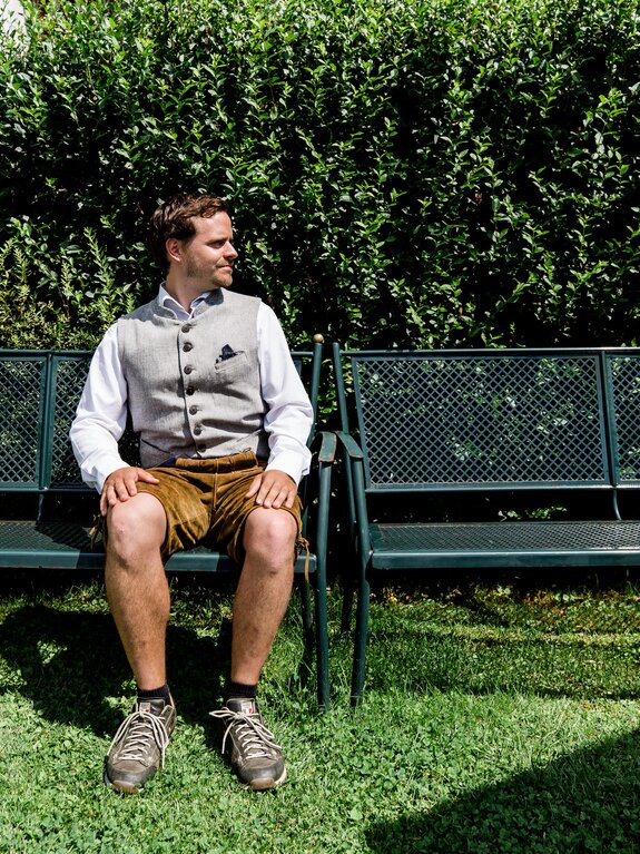 Gastgeber Andreas in Tracht sitzt auf Bank im Garten und schaut nach links