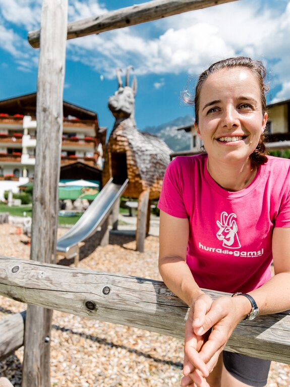 Tamara in pinkem T-Shirt lächelt auf Spielplatz mit Holzziege und Rutsche
