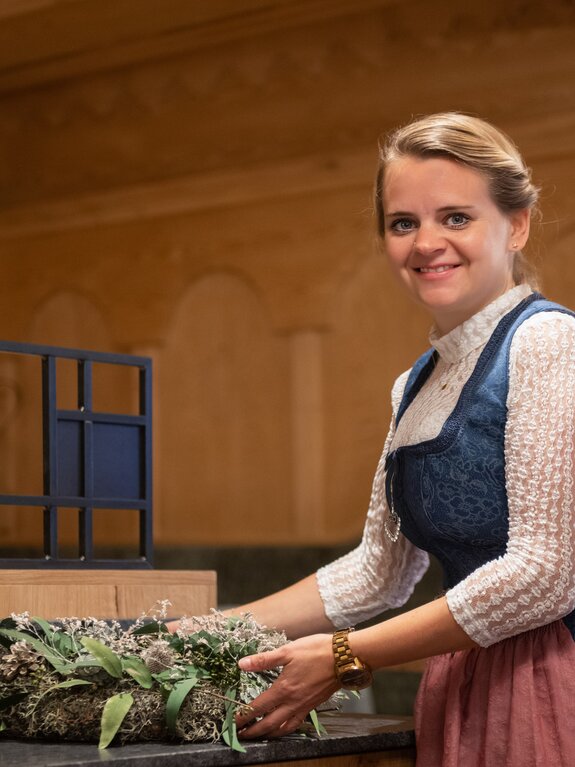 Frau im Dirndl arrangiert Blumenschmuck in einem holzvertäfelten Raum