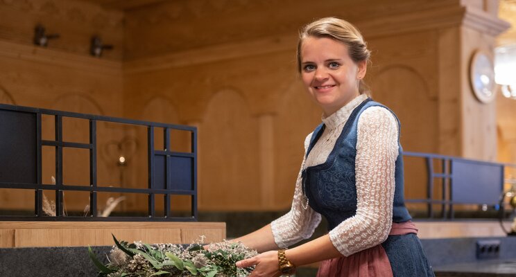 Frau im Dirndl arrangiert Blumenschmuck in einem holzvertäfelten Raum