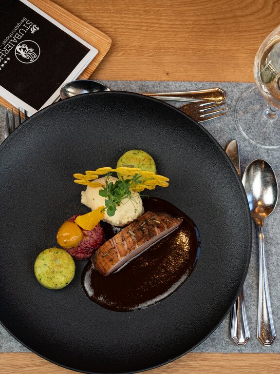 Teller mit Fleisch, Soße und Beilagen auf einem Tisch mit Besteck und Glas Wein