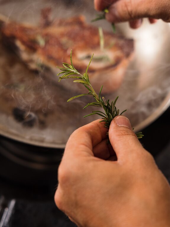 Hände halten Rosmarinzweig über dampfende Pfanne mit Fleisch