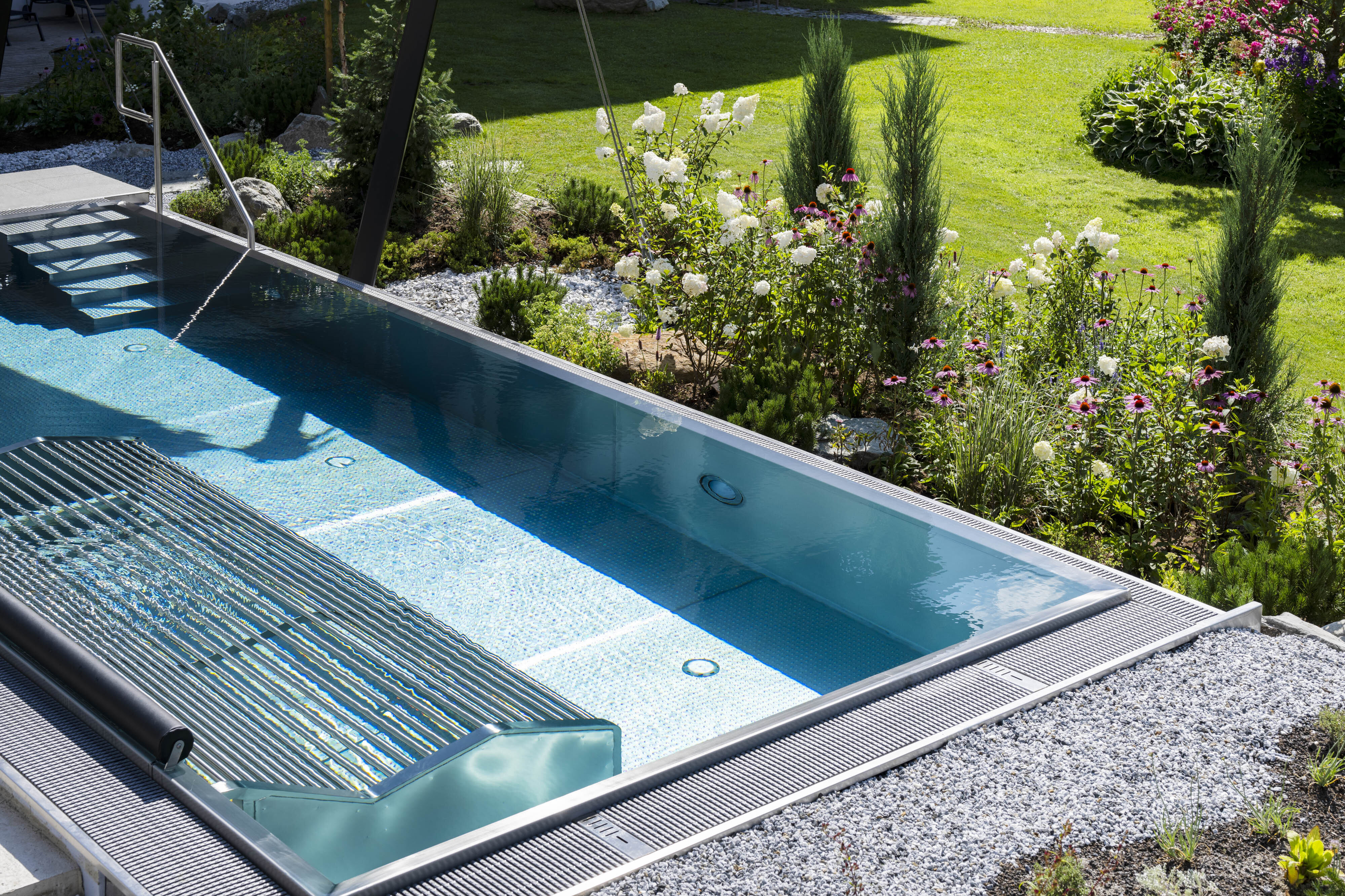 Rechteckiger Pool mit klarem Wasser, umgeben von Rasen, Blumen und Kies