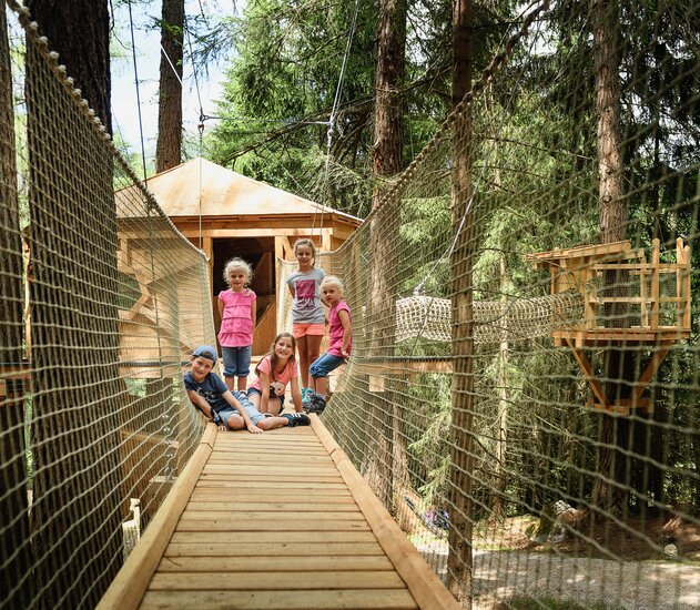 Fünf Kinder auf einer Hängebrücke vor einem Baumhaus im Wald