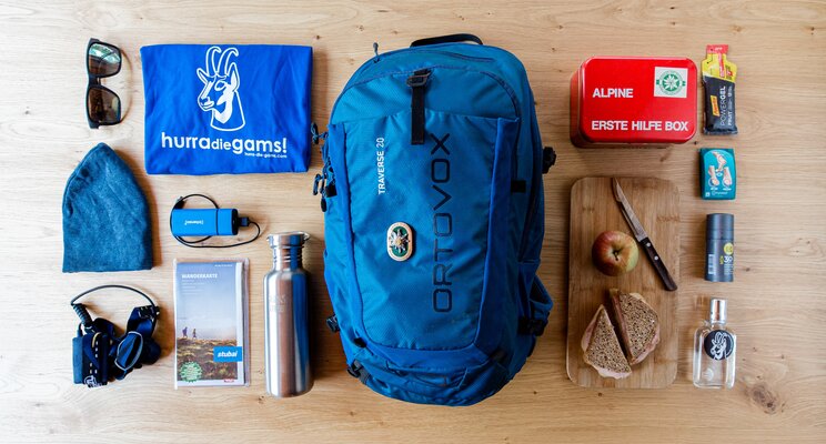 Blauer Rucksack, Erste-Hilfe-Kasten, Trinkflasche, Brotzeit und Wanderkarte auf Holztisch