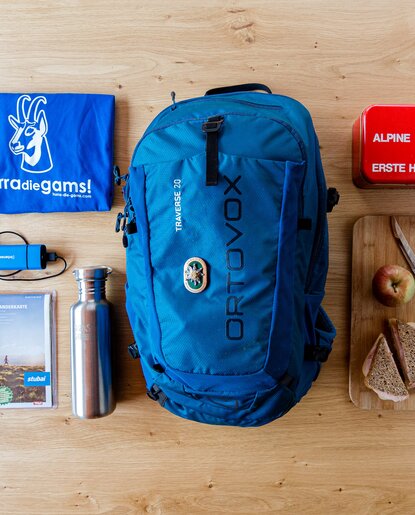 Blauer Rucksack, Erste-Hilfe-Kasten, Trinkflasche, Brotzeit und Wanderkarte auf Holztisch